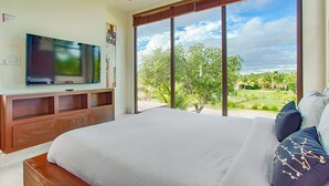 Un dormitorio con una cama grande, un televisor sobre un mueble de madera y vistas a un jardín a través de amplios ventanales.