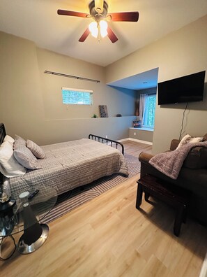 Un dormitorio con cama, ventilador de techo, televisión, ventana y una mesita.