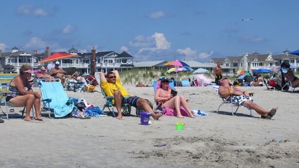 A beach scene with people relaxing on chairs and towels, umbrellas, and beach chairs scattered around.