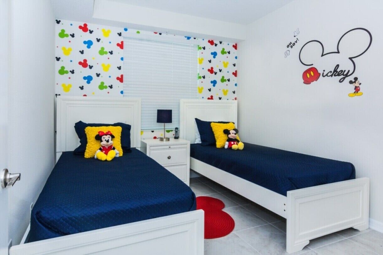 Een tweepersoonskamer met twee bedden, elk met een Mickey Mouse kussen, witte hoofdeinden en een klein nachtkastje met een lamp.