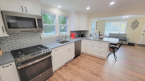 A modern kitchen with white cabinets, a stainless steel range, and a microwave. There is a dining area with a gray sofa and a small table.