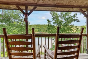 A wooden deck with two chairs facing a lake, surrounded by trees and a clear sky.