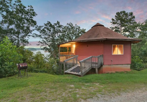 A small, single-story cabin with a porch and a staircase leading to a deck, surrounded by trees and a body of water.