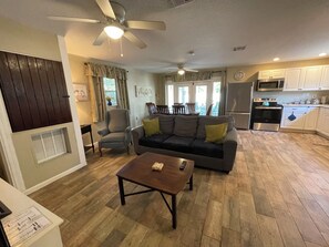 Ein Wohnzimmer mit einer Couch, Sesseln, einem Couchtisch und einem Deckenventilator. Es gibt eine Küchenzeile mit Geräten und Schränken.