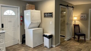 A laundry area with a washer and dryer, a trash bin, a wall-mounted sign, a door leading to a shower, and a chair. - ￼  Peaceful Two Bedroom Guest House near Wekiva River and State Parks ￼ (Sorrento)