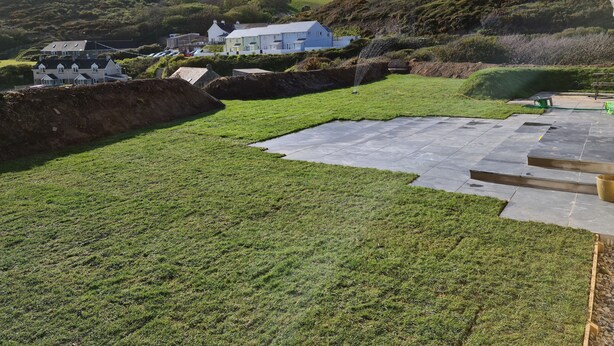 Un jardin aménagé avec une terrasse dallée, une pelouse et une colline en arrière-plan, sur laquelle se trouvent des maisons.