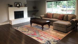 Una sala de estar con chimenea de ladrillo blanco, un sofá color beige con muchos cojines, una mesa de centro de madera y un tapiz colorido.