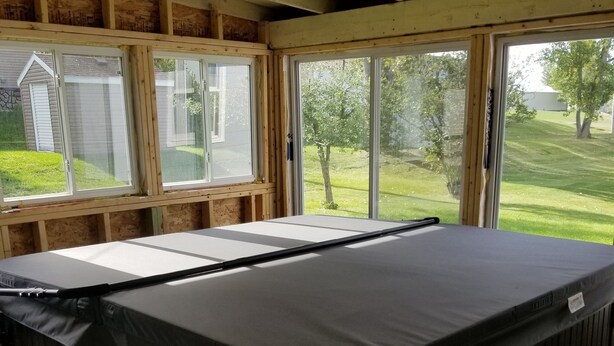 A hot tub in a room with large windows, wooden beams, and a view of a grassy yard.