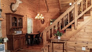 A wooden interior with a dining area, a chandelier, a staircase, and a mounted deer head.