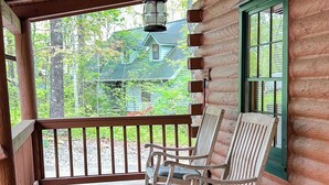 A wooden cabin with a porch featuring rocking chairs and a view of a green house in the background.