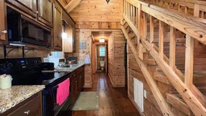 A wooden cabin interior with a kitchen, a staircase, and a bathroom visible through an open door.
