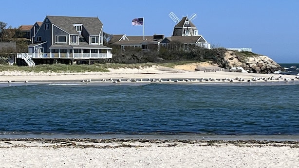 Una casa costera con un molino, una playa en primer plano y un cielo despejado.