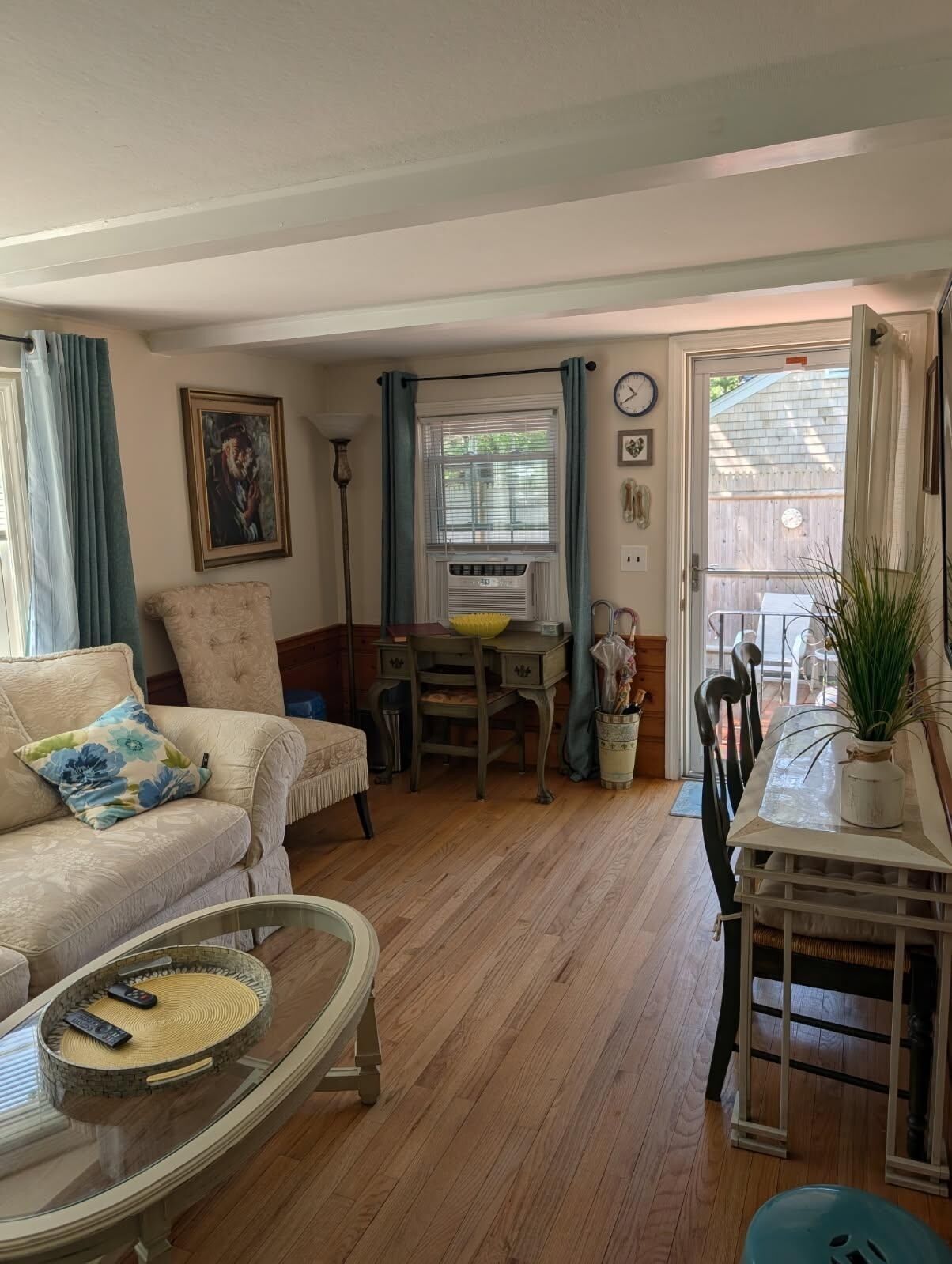 A cozy living room with a sofa, a glass coffee table, a dining table with chairs, a small desk, and a window with curtains.
