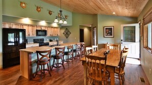 A kitchen with wooden cabinets, a large island with bar stools, a black refrigerator, and a hanging light fixture. - Sugar Bay Sweets - Lake Namakagon (Cable)