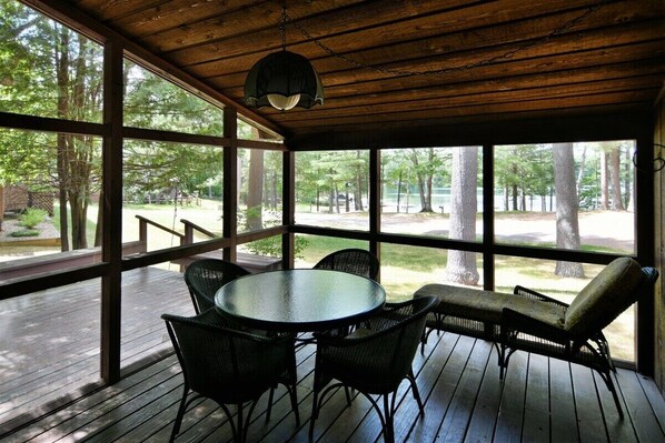 A covered outdoor area with a round table and chairs, large windows, and a view of trees.