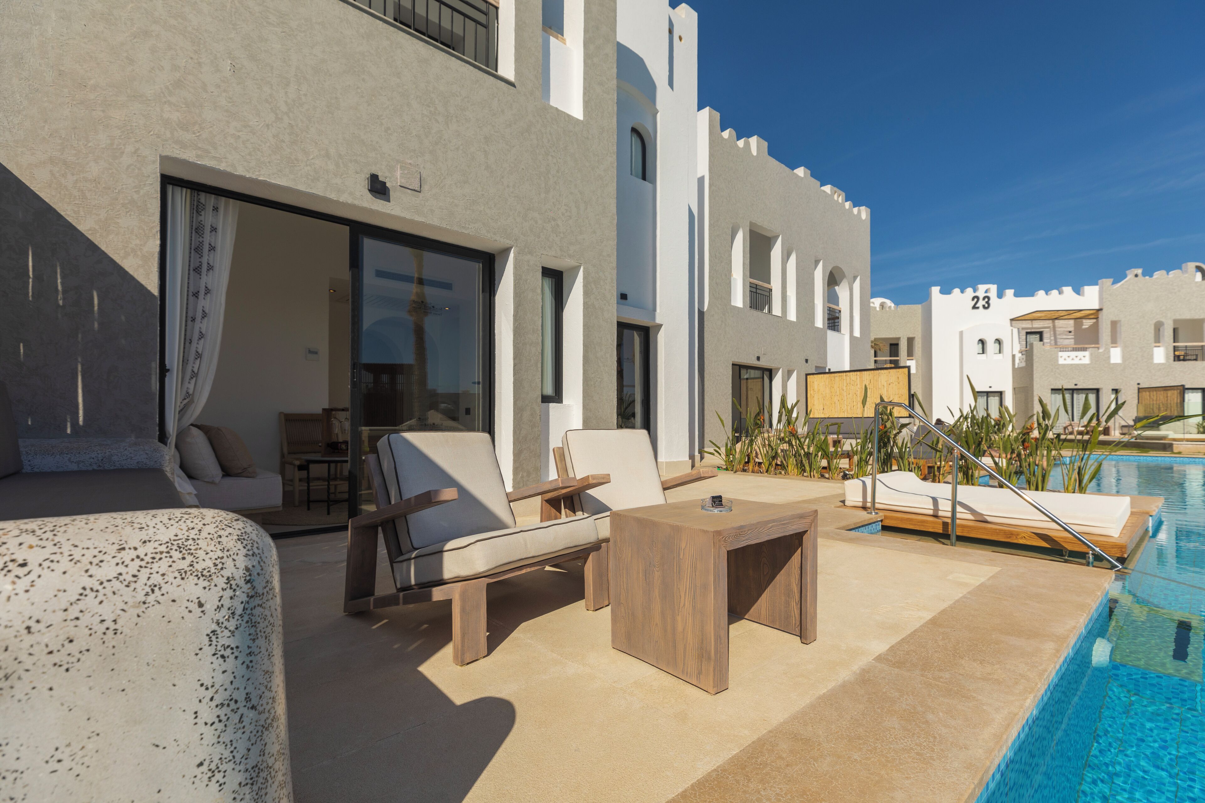 A modern outdoor pool area with lounge chairs and a wooden table, surrounded by a building with a white facade and numbered windows.