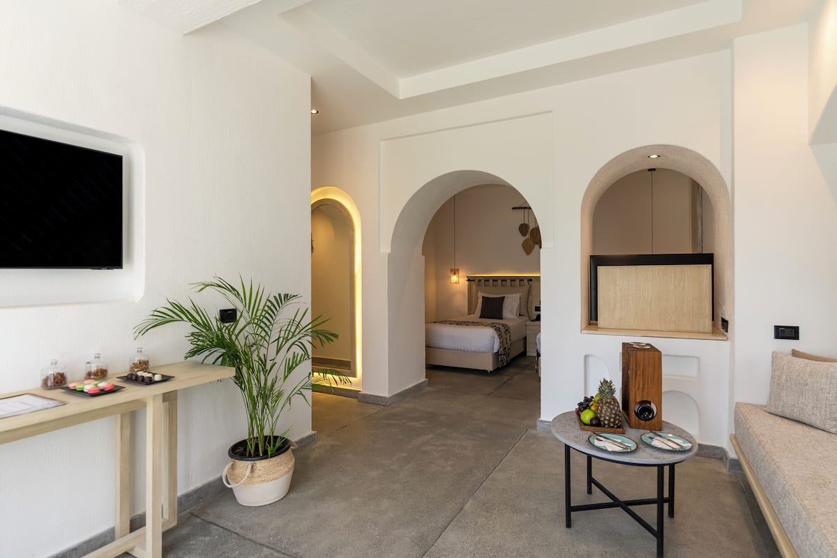 Une chambre d’hôtel moderne comprenant un lit, un bureau agrémenté d’une plante, une petite table avec un haut-parleur et une porte en arc donnant sur une autre pièce.