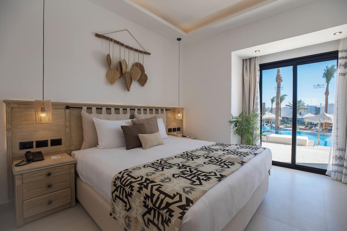 Une chambre d’hôtel avec un grand lit, des tables de chevet, un élément décoratif suspendu, et une vue sur une piscine et des palmiers.