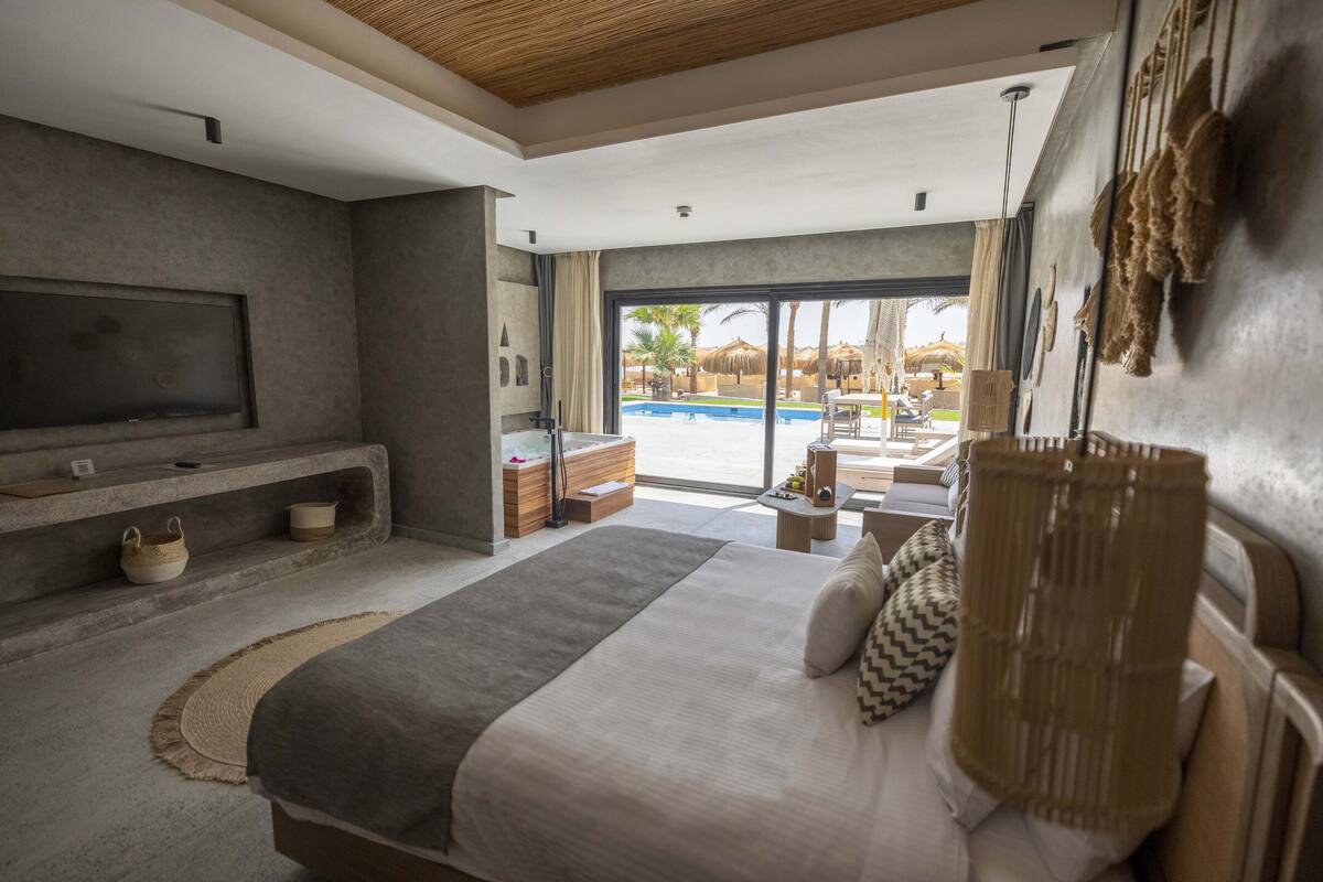 Une chambre moderne avec un grand lit, une télévision, une petite table et une vue sur une piscine et un espace extérieur.