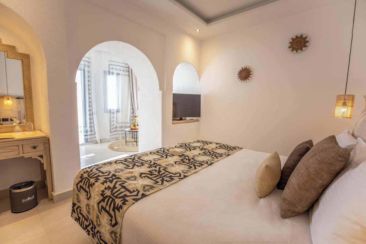 Une chambre avec un grand lit, un bureau et une chaise. Il y a une fenêtre avec des rideaux, un téléviseur fixé au mur et une décoration en forme de soleil.