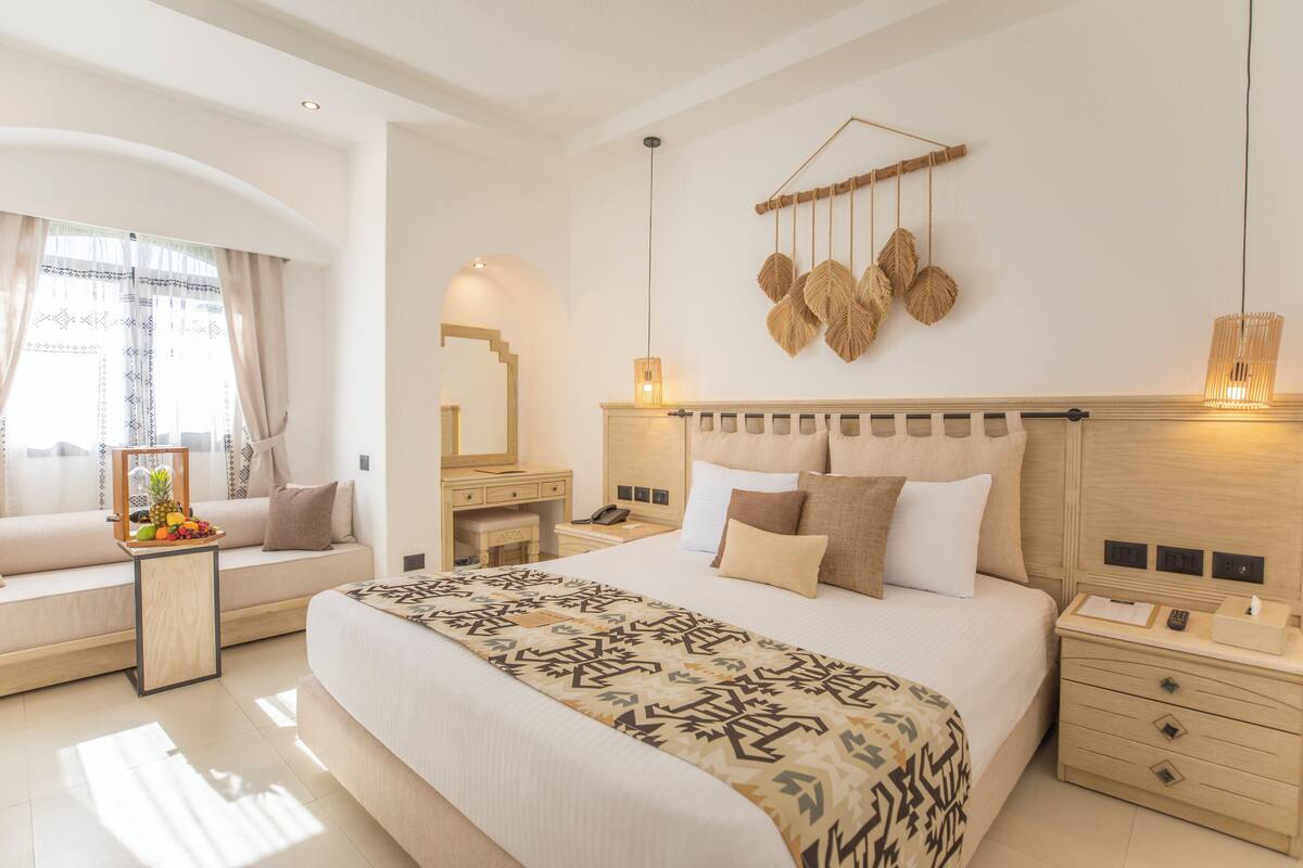 Une chambre d’hôtel moderne dotée d’un grand lit, de tables de chevet, d’une coiffeuse et d’un coin salon offrant une vue sur l’extérieur.