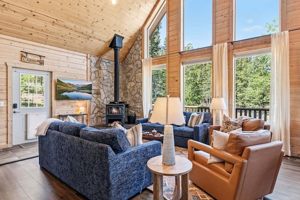 Ein Interieur einer Holzhütte mit Steinwand, Kamin, einer Couch, einem Sessel und einem großen Fenster.
