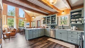 A spacious kitchen with wooden beams, a dining area with a sofa and table, and large windows offering a view of trees. - Custom Craftsman Chalet🏡Fenced Yard for Pets🍃Aspens, Hot Tub 💰$0 Cleaning & Pet Fee (Fairplay)