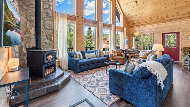 A cozy cabin interior with a stone fireplace, large windows, and a blue sofa. - Custom Craftsman Chalet🏡Fenced Yard for Pets🍃Aspens, Hot Tub 💰$0 Cleaning & Pet Fee (Fairplay)