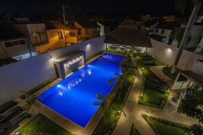 Vista nocturna de un resort con piscina, sillones de descanso y una estructura de techo de paja.