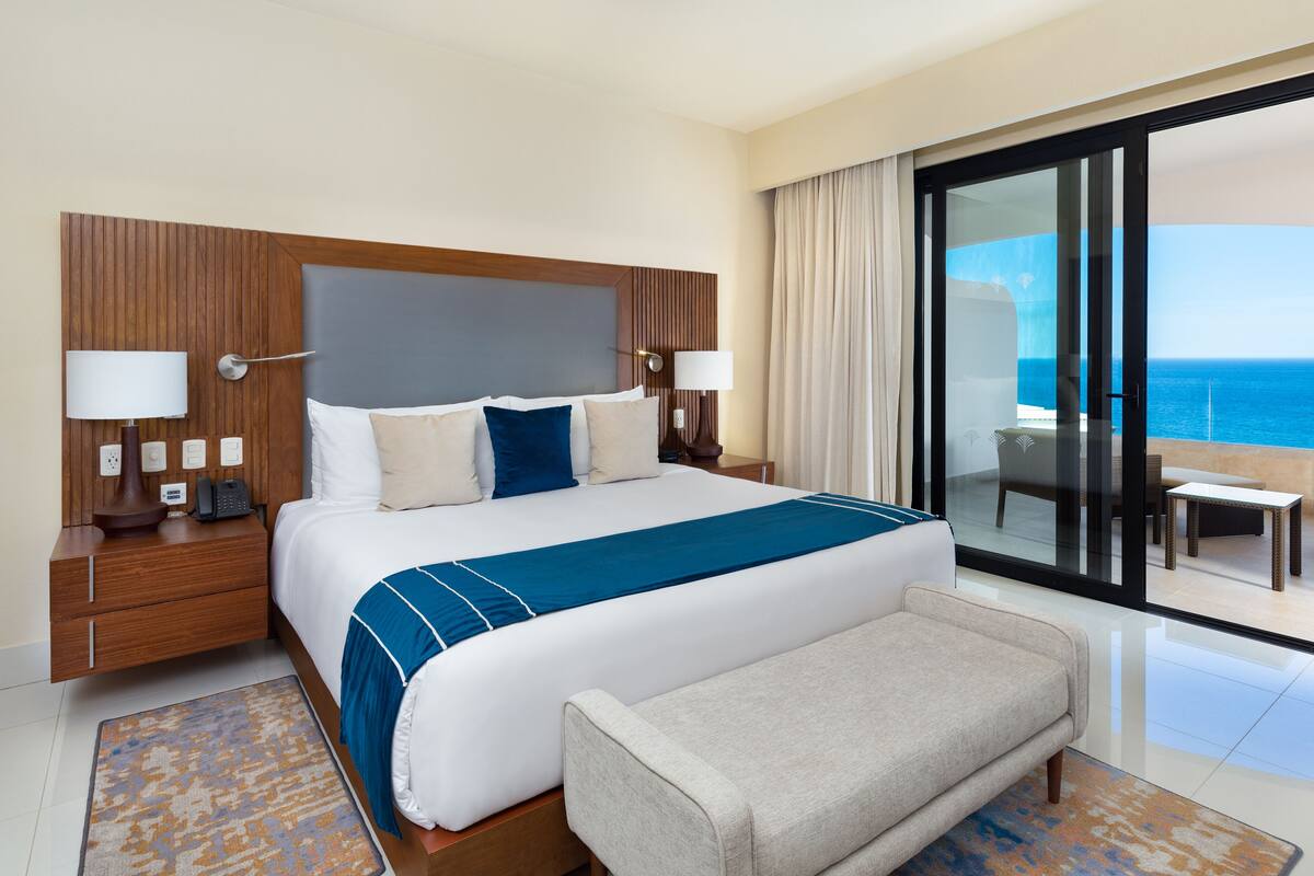 Ein Hotelzimmer mit einem großen Bett, einer Bank, einem Nachttisch mit Telefon und Blick auf das Meer durch eine Schiebetür.