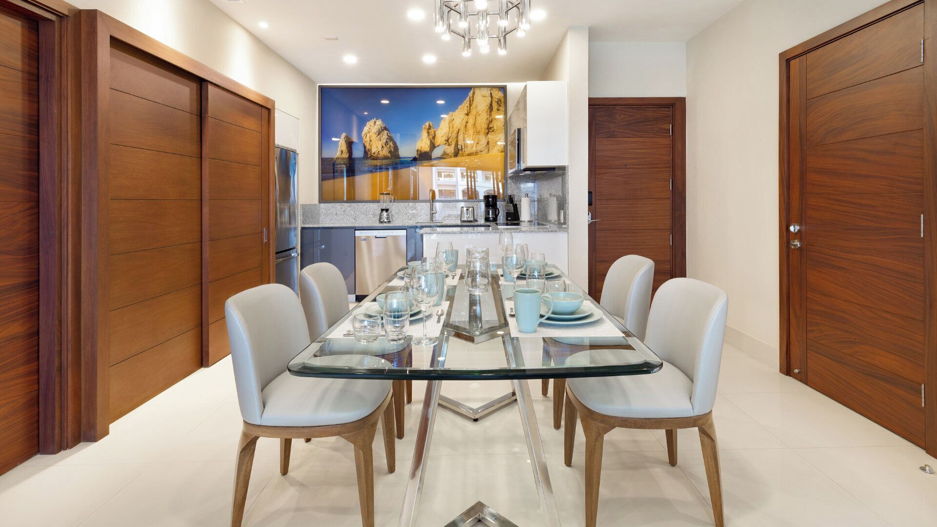 Un comedor con mesa de vidrio, sillas blancas y una cocina al fondo.