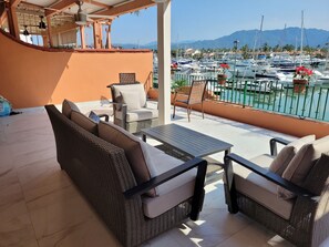 A balcony with wicker furniture, a table, and a view of a marina with boats and mountains in the background.