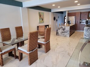 A dining area with wicker chairs and a glass table, a kitchen with a refrigerator and coffee maker, and a marble floor.