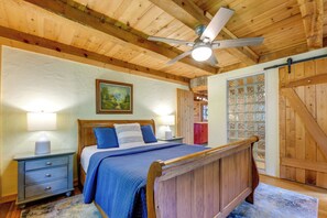 A bedroom with a wooden ceiling, a bed with blue bedding, a nightstand, a lamp, and a painting on the wall.