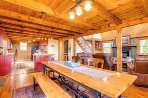 A spacious cabin interior with a wooden dining table, a kitchen area, a living room with leather furniture, and a staircase leading to an upper level.