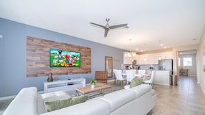 A modern living room with a large flat-screen TV, a wooden accent wall, a ceiling fan, and a dining area with white furniture.