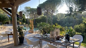 Un área de comedor al aire libre con una mesa dispuesta para dos personas, rodeada de sillas blancas, con una piscina y vegetación de fondo.