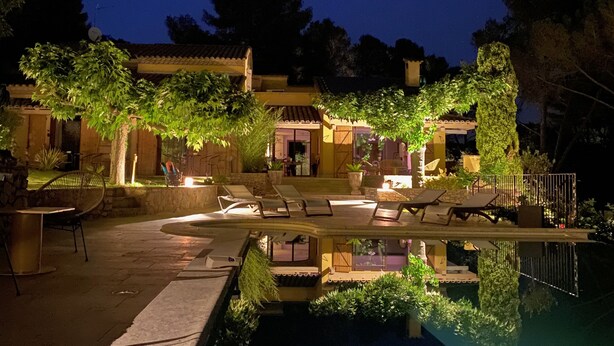 Vista nocturna de un área de piscina con sillones de descanso y una piscina cristalina que refleja el entorno.