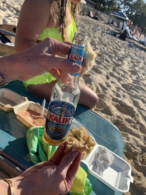 Una persona sosteniendo una botella de cerveza Kalik, con una playa y gente de fondo.