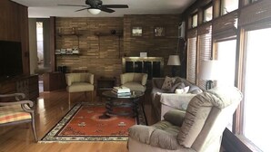Un salon doté d’un plancher en bois, d’un ventilateur de plafond, d’une cheminée et d’une grande fenêtre avec des stores.
