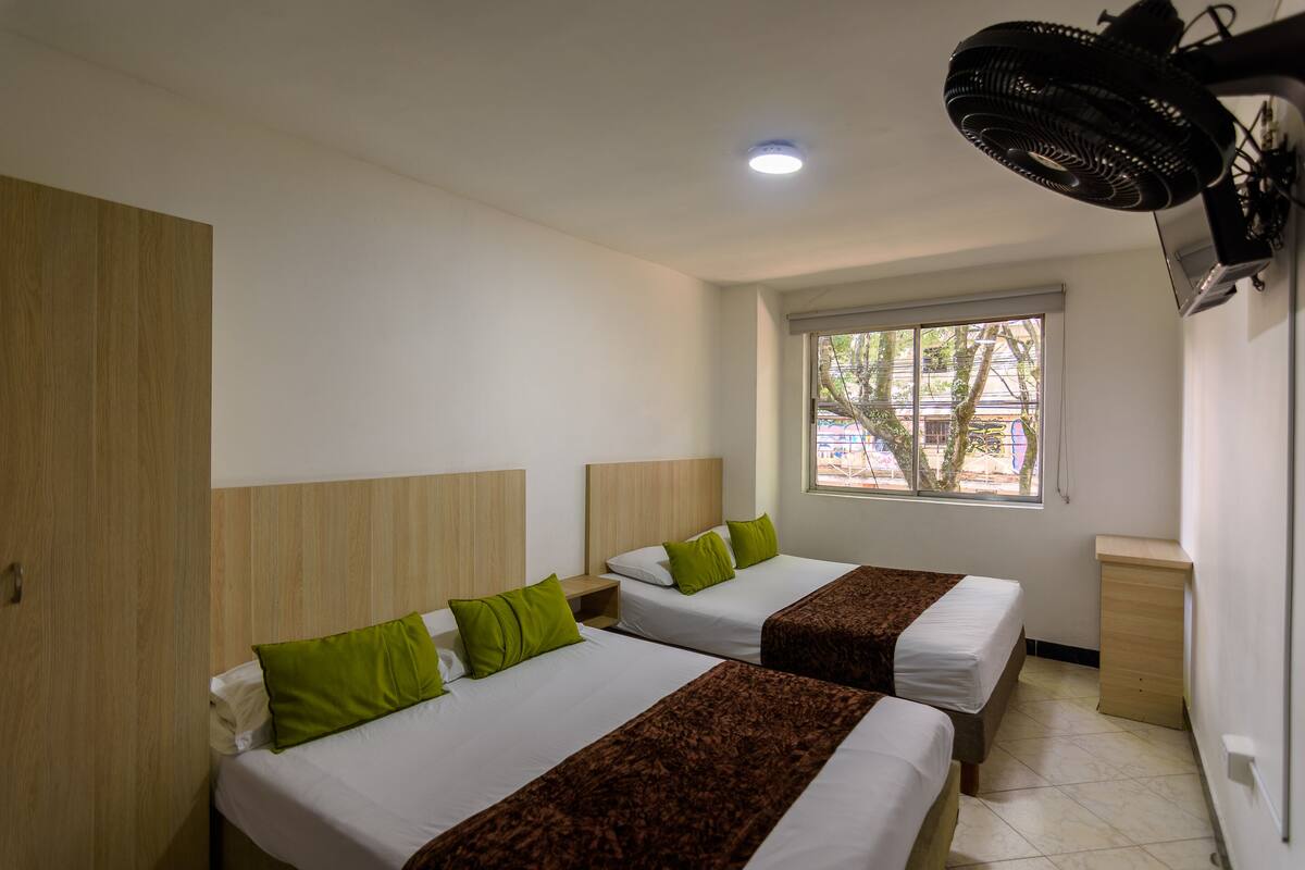 Una camera d'albergo con due letti, una finestra vista, un ventilatore a soffitto e un armadio in legno.