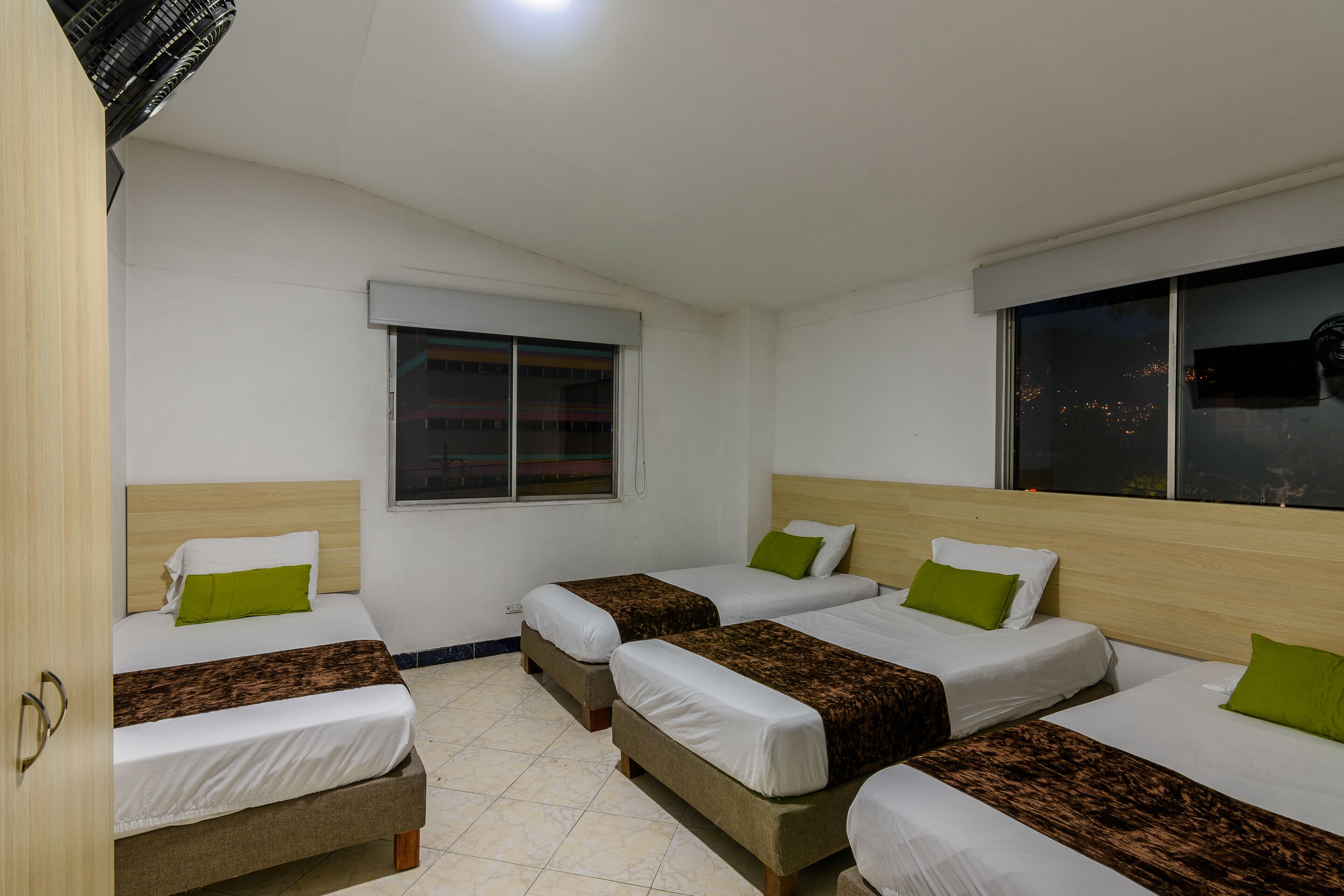 A hotel room with four beds arranged in a row, a window with a view of the city at night, and a door on the left side.