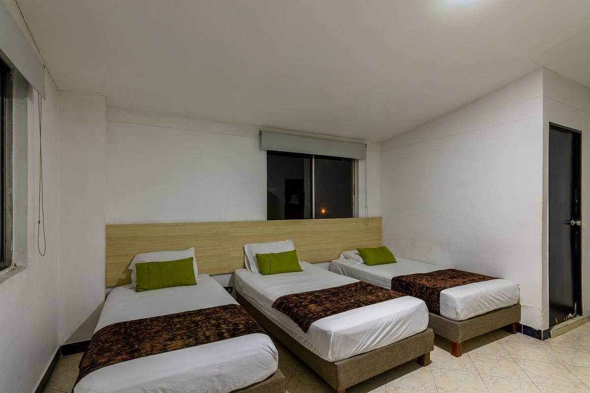 Camera d'albergo con tre letti singoli, ognuno con testiera in legno, biancheria da letto bianca e cuscini verdi. Si vede una finestra con vista all'esterno e una porta sulla destra.
