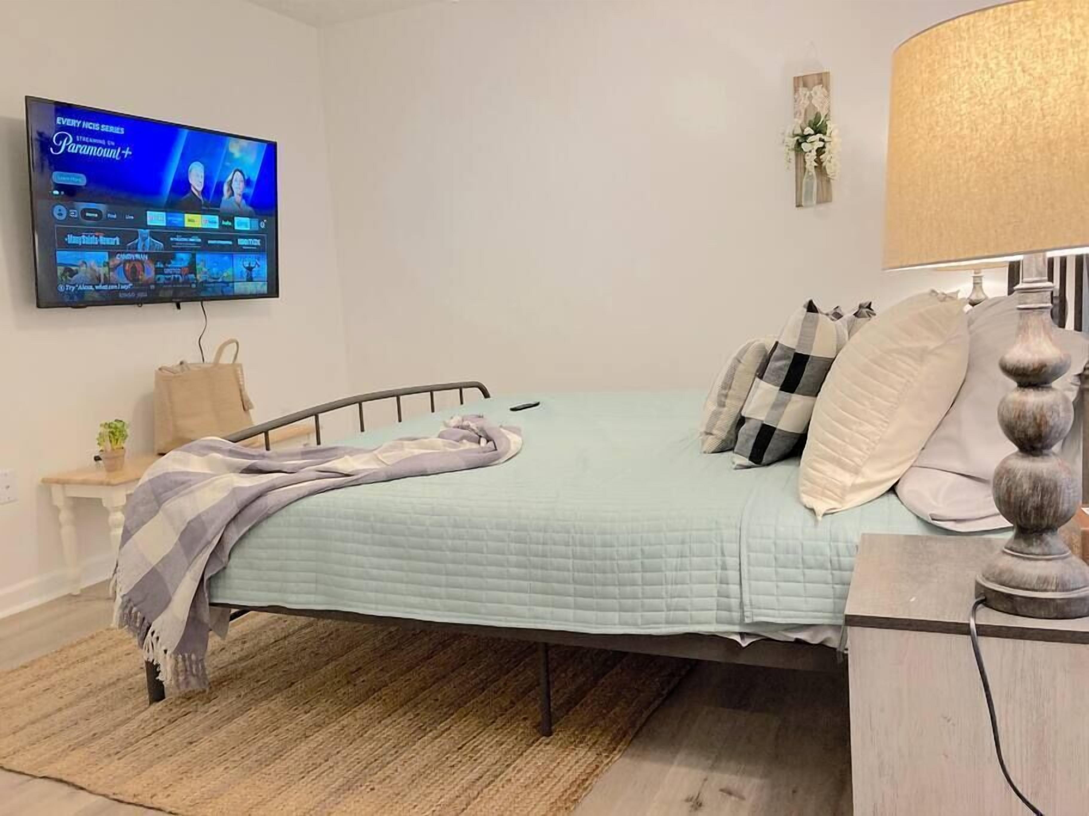 Ein Schlafzimmer mit einem Bett, einem an der Wand befestigten Fernseher, einem Nachttisch mit Lampe und einer kleinen Pflanze.