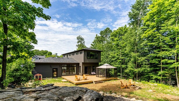 A modern two-story house with a deck, surrounded by dense forest.