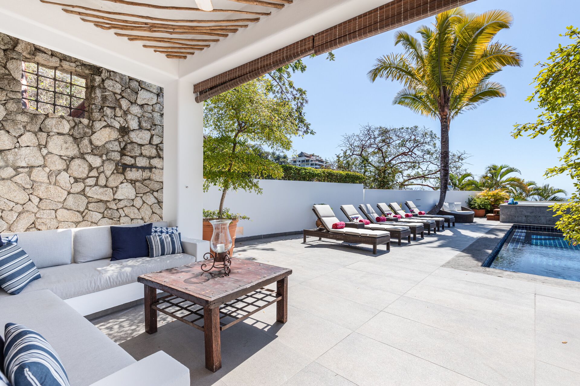 A rooftop patio with a stone wall, a pool, lounge chairs, and a table with a vase.