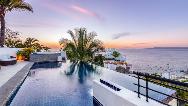 A infinity pool with a view of the sea at sunset, surrounded by palm trees and modern architecture.