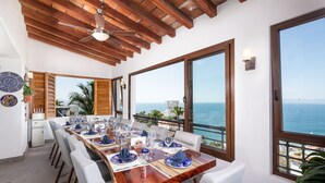A dining area with a wooden table set for six, large windows offering a view of the sea, and a ceiling fan.