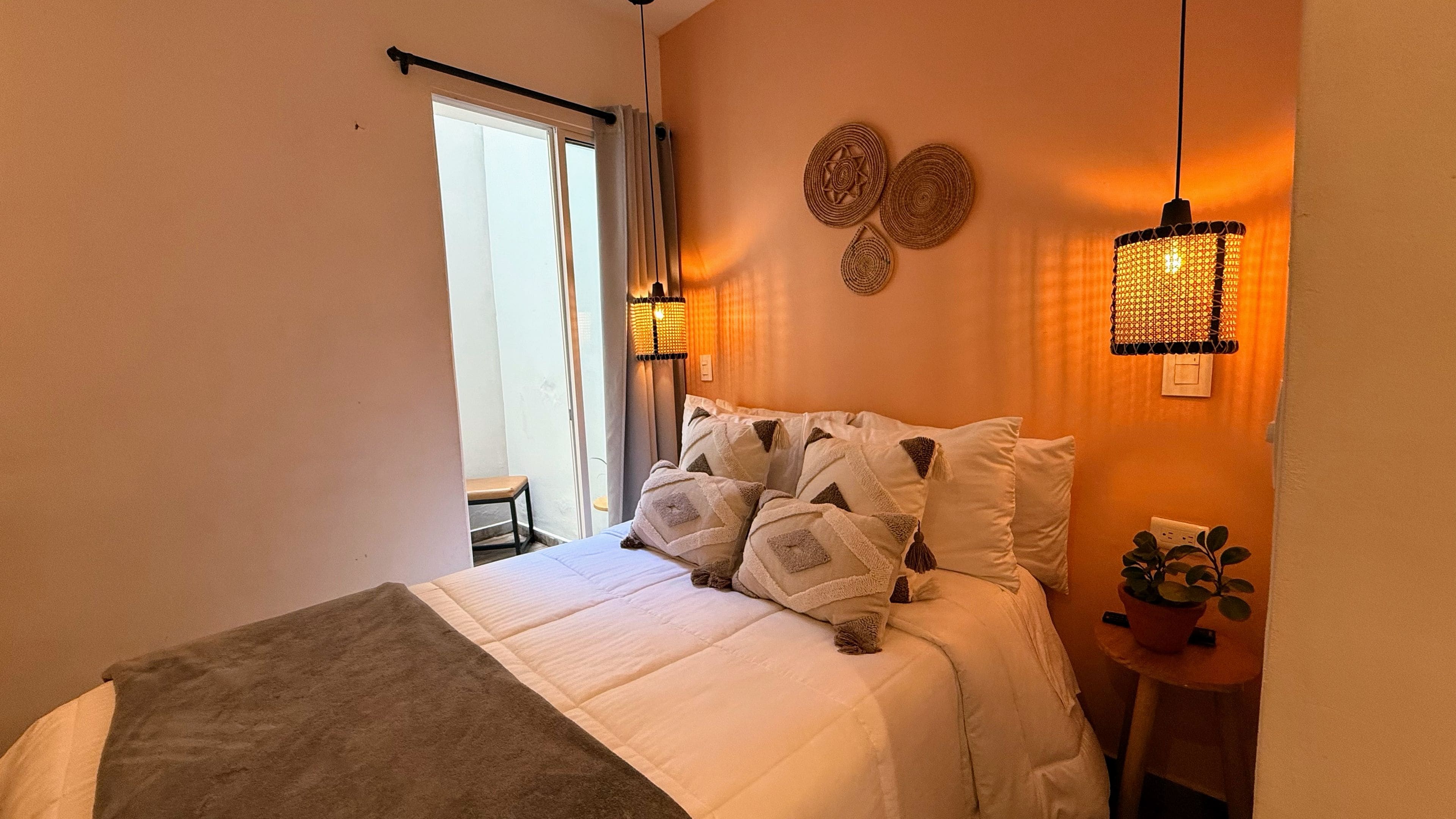Ein Schlafzimmer mit einem Bett, Kissen, einem Nachttisch mit Pflanze und einem Fenster mit Vorhängen.