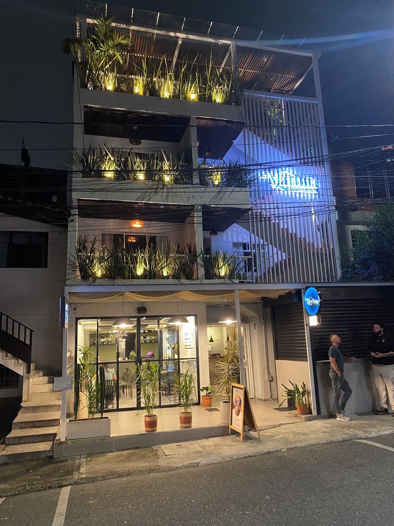 A two-story building with a balcony on the upper level, potted plants, and a sign with illuminated text.